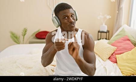 afroamerikanischer Mann in Kopfhörern, der spielerisch auf die Kamera in einem farbenfrohen Schlafzimmer zeigt Stockfoto