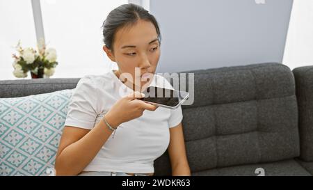 Junge asiatische Frau, die zu Hause eine Sprachnachricht auf dem Smartphone sendet und moderne Kommunikation in Innenräumen vorstellt. Stockfoto