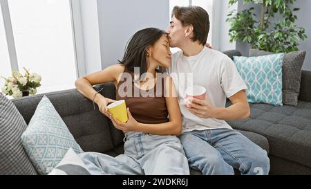 Interrassisches Paar, Mann, der Frau auf die Stirn küsst, Tassen hält, lässig gekleidet, entspannt auf Sofa in einem modernen Wohnzimmer, vermittelt Liebe und Togeth Stockfoto