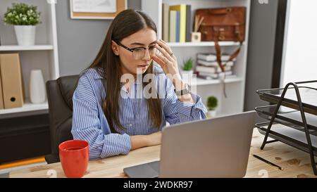 Gestresste junge hispanische Frau am Arbeitsplatz mit Laptop mit Brille. Stockfoto