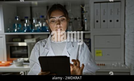 Hispanische Frau im weißen Labormantel untersucht eine Tablette in einem schwach beleuchteten Laborraum, umgeben von wissenschaftlichen Geräten. Stockfoto