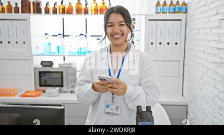 Eine lächelnde junge hispanische Frau in einem Labormantel benutzt ein Smartphone in einem klinischen Labor. Stockfoto