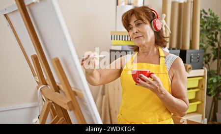 Eine ältere Frau malt auf einer Leinwand in einem Studio, trägt Kopfhörer und eine gelbe Schürze und hält einen roten Becher. Stockfoto