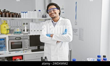 Selbstbewusster Wissenschaftler mit lockigen Haaren, die in einem Labor lächeln, mit Laborkittel und Schutzbrille und überkreuzten Armen Stockfoto