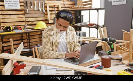 Ein afroamerikaner arbeitet an einem Laptop in einer gut ausgestatteten Tischlerei, umgeben von Werkzeugen und Holzstücken. Stockfoto