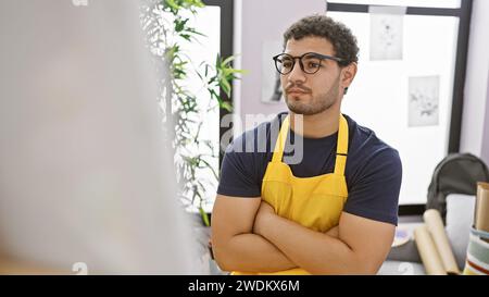 Ein selbstbewusster junger Mann in Brille und gelber Schürze steht in einem kreativen Studio mit Kunstwerken überkreuzt. Stockfoto