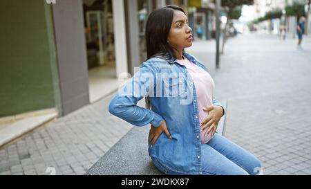 Müde junge schwangere Frau, die ihren Bauch berührt, sitzt entspannt auf einer städtischen Straßenbank und gleicht die Emotionen einer werdenden Mutterschaft aus Stockfoto