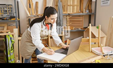 Junge hispanische Frau, die in der Werkstatt mit Schreinerwerkzeugen und Holzkonstruktionen am Laptop arbeitet Stockfoto