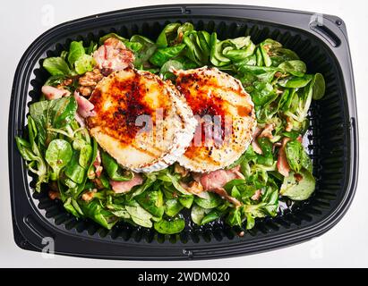 Gegrillter Ziegenkäse auf einem Bett aus frischem Spinat und Zucchini mit Walnüssen in einem schwarzen Plastikbehälter, bereit zum Mitnehmen. Stockfoto