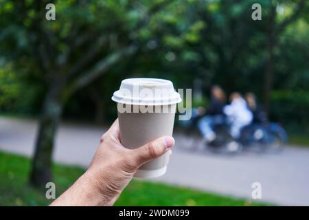 Die Hand eines Mannes hält eine Kaffeetasse mit Menschen, die draußen im Hintergrund verschwimmen. Stockfoto