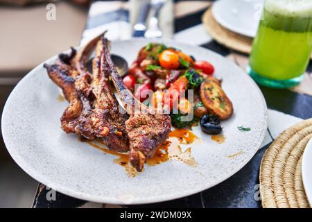 Gegrillte Lammkoteletts auf einer Keramikplatte mit Gemüse und grünem Smoothie in entspannter Atmosphäre im Freien. Stockfoto