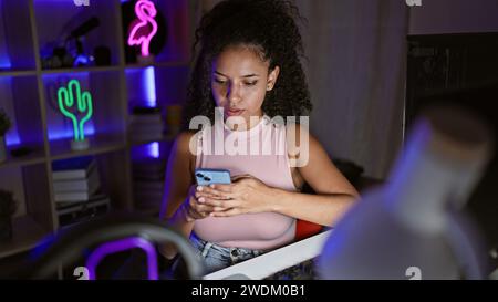 Attraktive junge hispanische Frau Streamer, vertieft in einen Gaming-Stream, beugte sich über das Smartphone in einem neonbeleuchteten Spielzimmer und strahlte eine entspannte und dennoch entspannte Atmosphäre aus Stockfoto