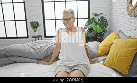 Selbstbewusste, grauhaarige Seniorin, mit kurzen Haaren und Brillen, die einen gemütlichen Morgen in ihrem Zuhause genießt. Bequem sitzen, auf dem Bett ruhen Stockfoto