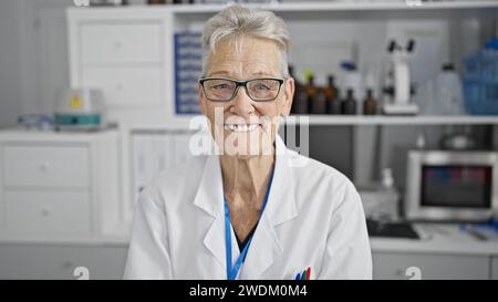 Selbstbewusste und lächelnde grauhaarige Wissenschaftlerin genießt ihre Arbeit und sitzt glücklich im Labor zwischen Mikroskop und Reagenzgläsern Stockfoto