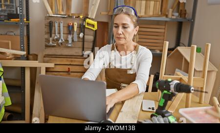 Eine reife Frau in einem Holzverarbeitungsstudio konzentriert sich auf ihren Laptop, inmitten verschiedener Zimmermannswerkzeuge. Stockfoto