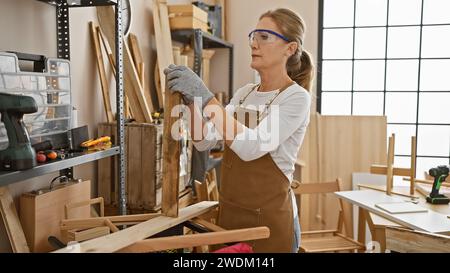 Handwerkerin mittleren Alters inspiziert Holz in sonnigen Werkstätten Stockfoto