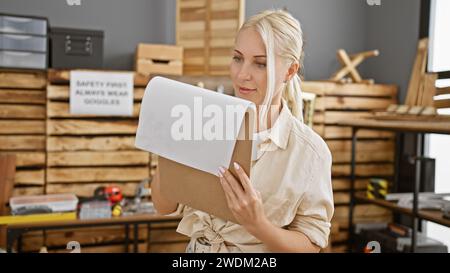 Die attraktive junge blonde Tischlerin macht fleißige Notizen auf die Zwischenablage während der Holzarbeiten in der geschäftigen Tischlerei Stockfoto