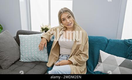 Fröhliche junge blonde Frau, die zu Hause auf dem Sofa sitzt, mit strahlendem Lächeln sprach und voller Selbstvertrauen war Stockfoto