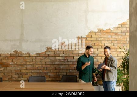 Zwei Profis führen ein fokussiertes Gespräch, während sie ein digitales Tablet in einem modernen Bürobereich mit rustikalem, freiliegendem Mauerwerk halten Stockfoto