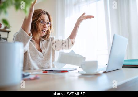 Schöne Frau mittleren Alters in Brille, die extrem überrascht war, dass sie neue höhere Steuern beobachtete und Rechnungen auf Laptop im Heimbüro warf. Kleine Unternehmen Stockfoto