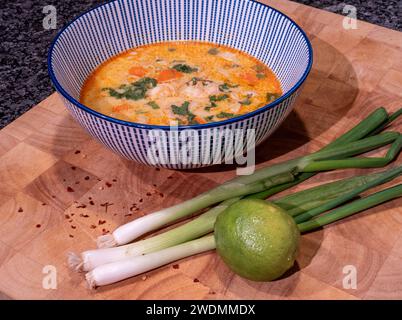 Genießen Sie die reichen Aromen einer pikanten asiatischen Kokossuppe mit roter Currypaste, die elegant in einer blauen Schüssel auf einem Holzblock präsentiert wird. Begleitet Stockfoto