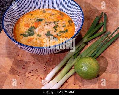 Genießen Sie die reichen Aromen einer pikanten asiatischen Kokossuppe mit roter Currypaste, die elegant in einer blauen Schüssel auf einem Holzblock präsentiert wird. Begleitet Stockfoto