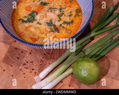 Genießen Sie die reichen Aromen einer pikanten asiatischen Kokossuppe mit roter Currypaste, die elegant in einer blauen Schüssel auf einem Holzblock präsentiert wird. Begleitet Stockfoto