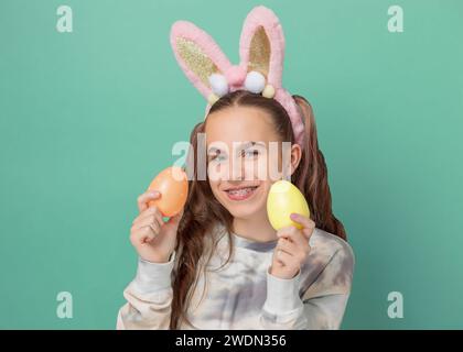 Frohe Ostern. Lustiges süßes Mädchen mit Osterhasenohren und bunten Ostereiern auf grünem Hintergrund. Kopierbereich. Lächelndes Mädchen in Zahnspangen. Stockfoto