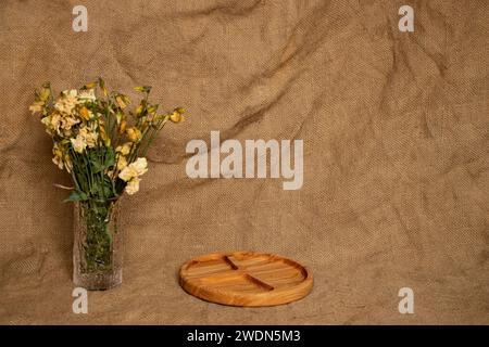 Leerer portionierter Holzteller, Vase mit getrockneten Blumen auf Sackleinen Hintergrund, minimalistischer Hintergrund zur Anzeige Stockfoto