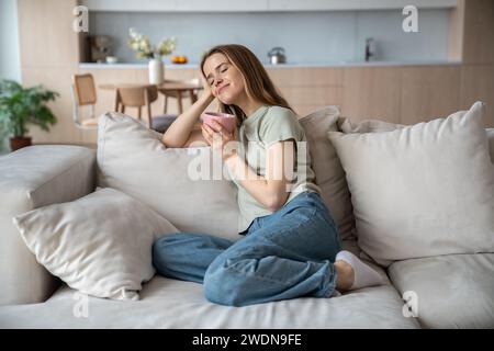 Fröhliche Frau, die auf dem Sofa sitzt, Tee trinkt, Freude, Zufriedenheit, Entspannung Stockfoto