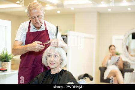 Der erfahrene Senior man Master macht Haarschnitt für reife Frauen mit einer Schere Stockfoto