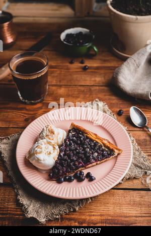 Ein Stück frische hausgemachte Amelanchier-Beeren-Galette und zwei Kugeln Vanille-Eiscreme auf dem Teller mit einem Glas Kaffee auf dem rustikalen Tisch Stockfoto