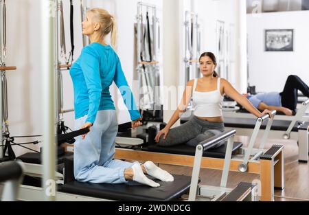 Zwei sportlich fokussierte junge Frauen in Sportbekleidung, die Pilates-Übungen für den Rücken auf einer reformierten Bettmaschine im Fitnessstudio durchführen Stockfoto
