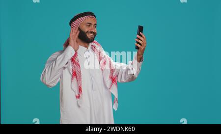 Muslimischer Erwachsener führt Online-Chat am Telefon in traditioneller islamischer Kleidung. Junge Person mit arabischem Kopfschmuck und du plauderst auf Videokonferenz mit Freunden im Studio. Stockfoto