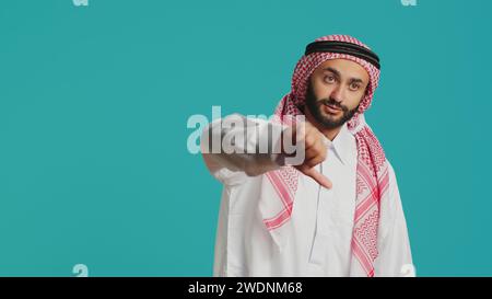 Muslimischer Kerl mit antikem Kleid und Kufiyah gibt die Daumen nach unten und fühlt sich mit Sachen unzufrieden. Junge Person drückt eine scheinbare Verleugnung und ein negatives Zeichen im Atelier aus, arabisches Modell. Stockfoto