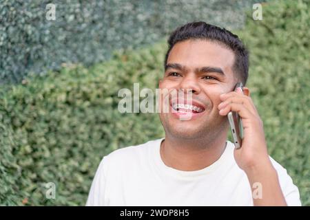 Ein lächelnder junger Mann mit Zahnspangen, der ein Gespräch auf seinem Smartphone genießt, draußen mit einer grünen Hecke im Hintergrund. Stockfoto