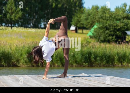 Schlanke Frau, die Yoga praktiziert, die Ardha Chandrasana-Übung, eine Variante der sichelförmigen Haltung, Training an einem Sommermorgen auf einem hölzernen p Stockfoto