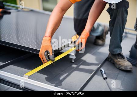 Arbeiter bauen Solarpaneelanlage auf dem Dach des Hauses zur Stromerzeugung durch Photovoltaik-Effekt. Nahaufnahme eines Mannes mit einem Lineal zur Messung der Montageausrüstung für eine präzise Installation. Stockfoto