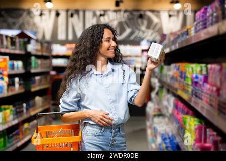 Fröhliche junge Frau mit lockigem Haar wählt Produkte im Supermarkt aus, die beispielhaft für gesunde Lebensweise und Einkaufsgewohnheiten sind. Stockfoto
