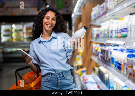 Lächelnde Frau mit lockigem Haar, die Haushaltsartikel kauft, eine Liste auf dem Smartphone im Supermarktgang überprüft. Stockfoto