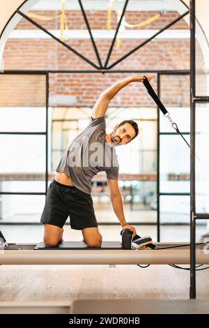 Junger bärtiger männlicher Sportler in Sportbekleidung, der in die Kamera blickt, während er auf dem Reformbett kniet und in einem modernen Fitnessstudio mit Pilates-Geräten trainiert Stockfoto