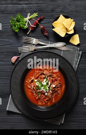 Birria de res, mexikanischer Rindereintopf in heißer Paprikasauce mit roher Zwiebeln und gehacktem Koriander, Taco-Chips in schwarzer Schüssel auf schwarzem Holztisch mit Gabel Stockfoto