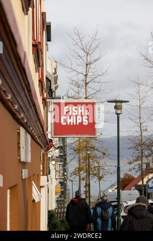 Rückansicht der anonymen Fußgänger in warmer Kleidung, während sie auf dem Bürgersteig in der Nähe des Gebäudes mit Restaurant, das Fish and Chips vor bedecktem Himmel serviert, spazieren gehen Stockfoto