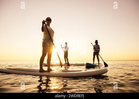 Drei aktive junge Touristen laufen auf Stand-up-Paddelbrettern am Sonnenuntergang See Stockfoto