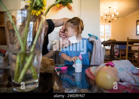 Mom wischt sich Töchtern das Gesicht mit Taschentüchern ab, während sie mit Spielzeug spielt Stockfoto