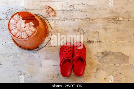 Auf weiß getünchten Dielen ein umgedrehter orangener Kunststoffeimer mit einem riesigen Loch im Boden und ein Paar knallroter Krokodile Stockfoto