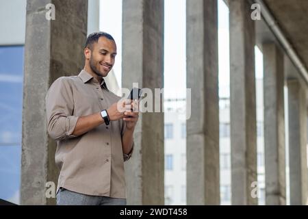 Lächelnder afroamerikanischer Geschäftsmann in lässigem Hemd, der draußen telefoniert, möglicherweise SMS oder Browsing. Stockfoto