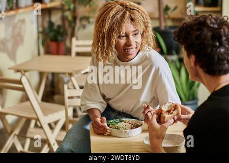 Glückliche afroamerikanerin mit Zahnspange, die einen lockigen Freund ansieht, der im Café veganen Burger isst Stockfoto