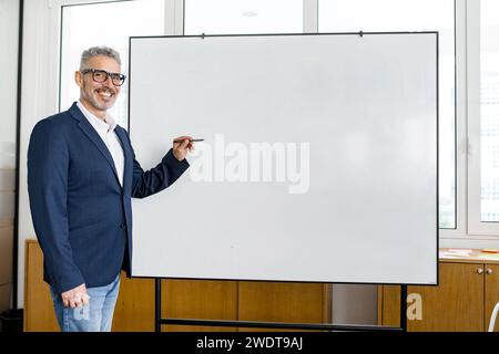Reifer, grauhaariger Geschäftscoach, der drinnen auf Whiteboard schreibt. Leitender Manager ceo Büromitarbeiter zeigt auf ein leeres Flipchart und schaut in die Kamera, hält Briefing, Schulung, Kopierraum ab Stockfoto
