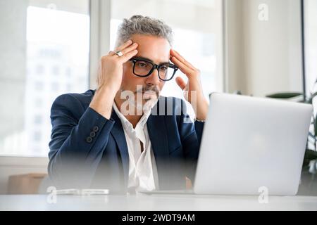 Ein reifer Geschäftsmann mit besorgtem Ausdruck massiert seine Schläfen, während er auf den Laptop-Bildschirm blickt, was auf eine tiefe Fokussierung oder Stress durch die Arbeit hindeutet, stellt eine anspruchsvolle Art von Unternehmensentscheidungen dar Stockfoto